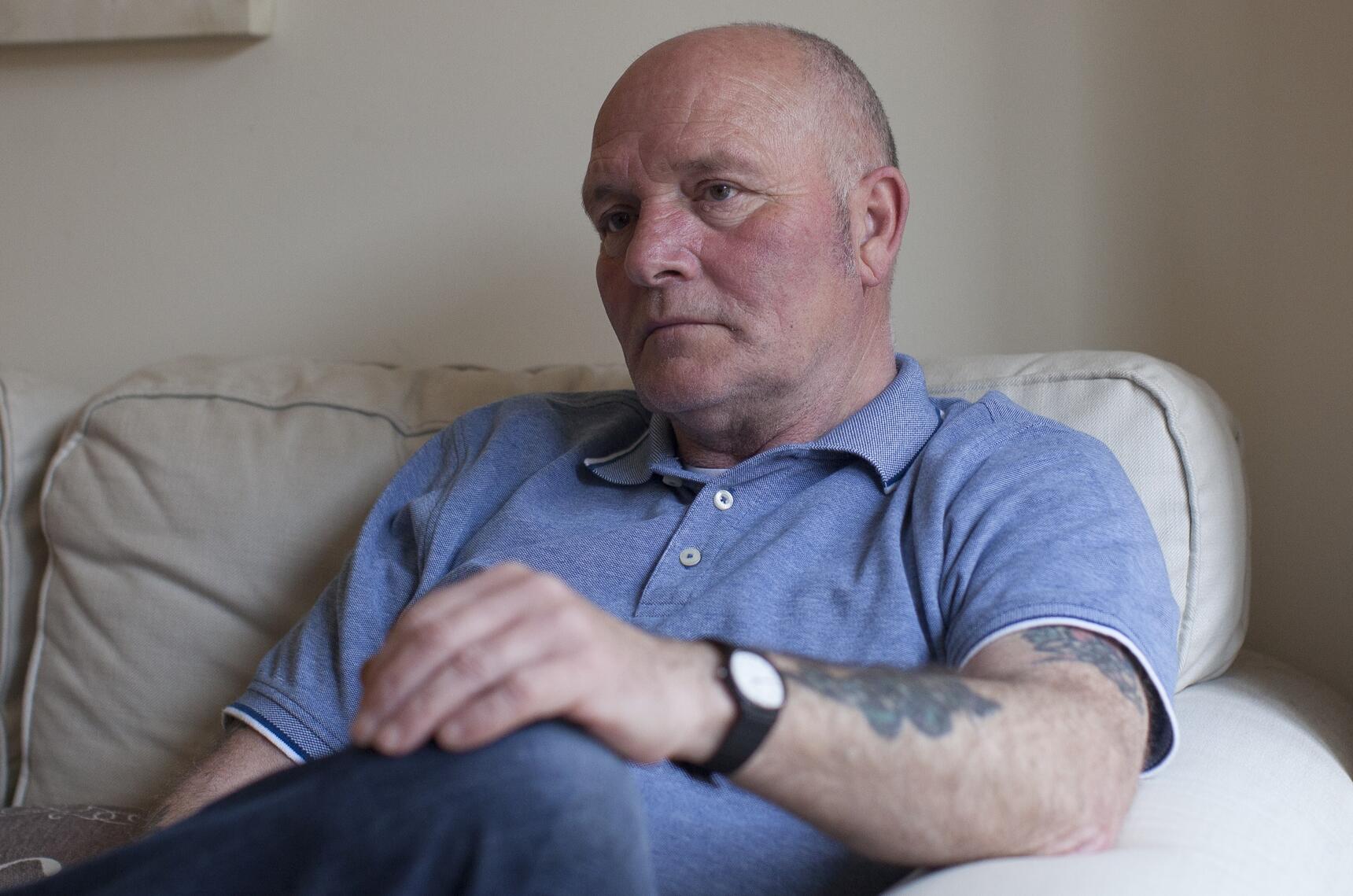 A bald man with a light blue top sat on a cream sofa