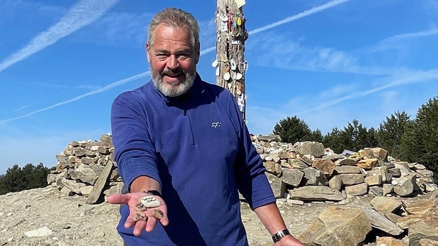 Kevin is holding out his hand with stones in his palm, he's wearing a blue top and behind him is monument