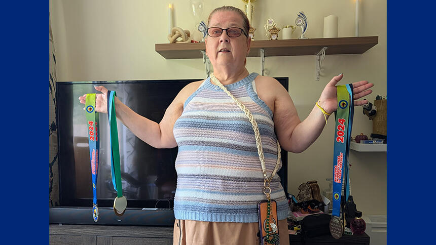 Trish is stood with her arms outstretched with all her archery medals, she is indoors and wearing a stripey blue top.