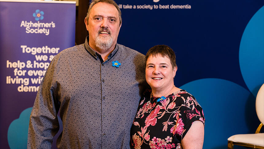 Julie is wearing a black and pink flowery dress, she's stood next to her husband David who is wearing a grey shirt. There is an Alzheimer's Society banner behind them.