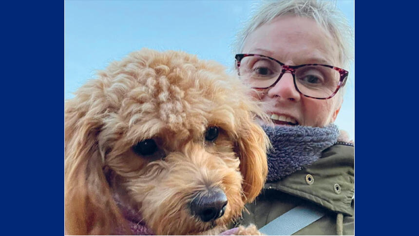 Sue has cropped grey hair and glasses, she's holding her dog Lily who has fawn-coloured fur.