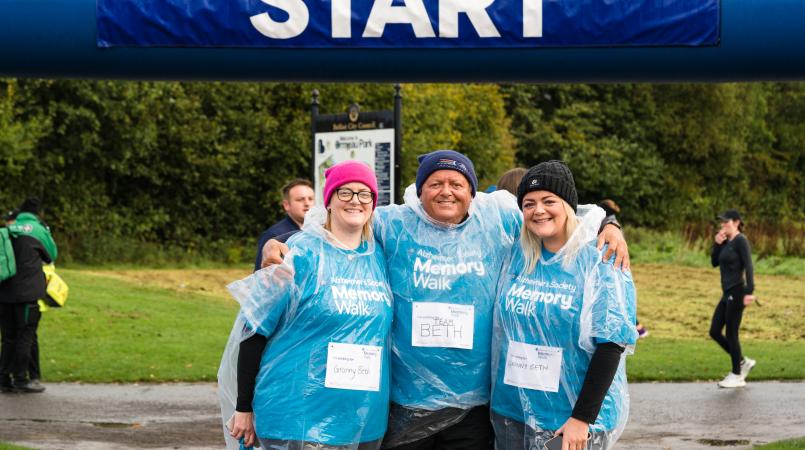 3 people in Memory Walk t-shirts and ponchos, smiling in front of the start line.