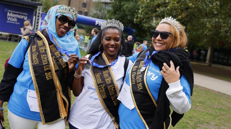 3 women in blue Memory Walk t-shirts, smiling at the camera wearing sashes and tiaras