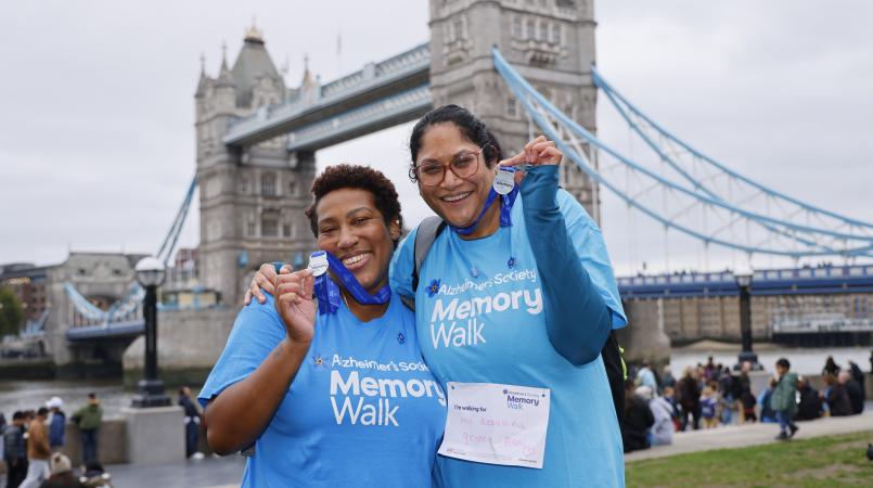 2 women in blue Memory Walk t-shirts smiling in front of Tower Bridge holding up their medals