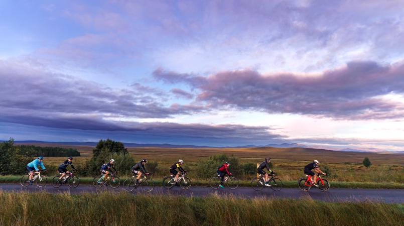 Cyclists taking part in event with landscape backdrop, cycling in a line with the sunset in the sky
