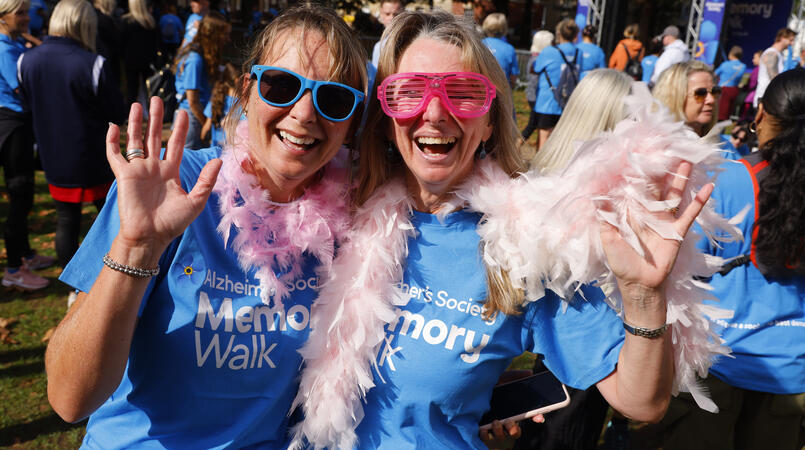 Two walkers smile and wave wearing blue Memory Walk t-shirts, sunglasses and pink feather boas
