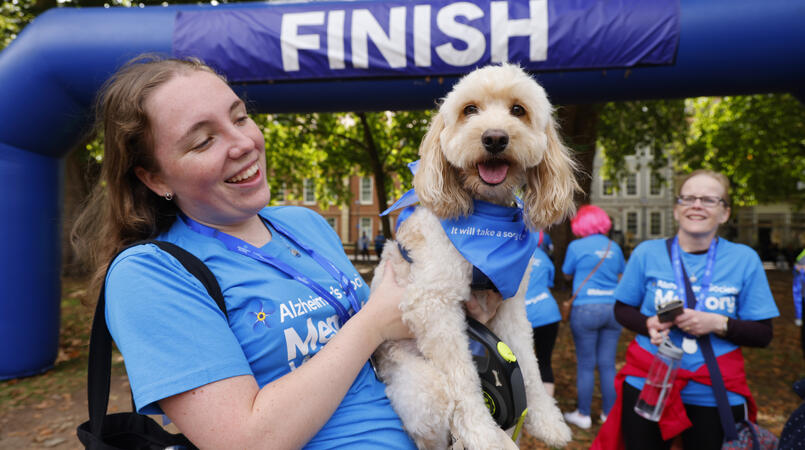 A walker in a blue Memory Walk t-shirt is holding their dog wearing a blue bandana at the finish line