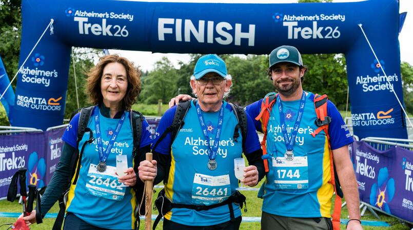 3 people with medals stood in front of a trek26 finish line.